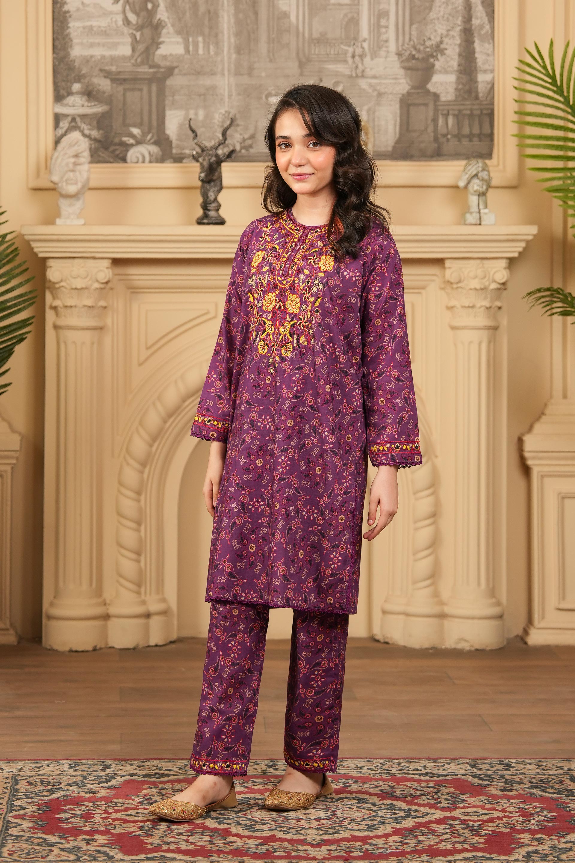 Woman wearing a purple embroidered outfit standing in a decorated room with a fireplace.