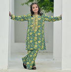 Woman in a green floral outfit standing in front of a white archway with greenery outside.