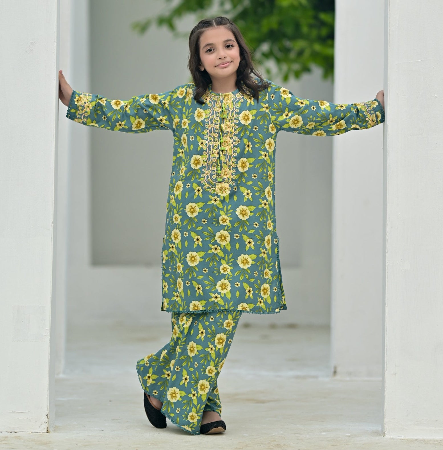 Woman in a green floral outfit standing in front of a white archway with greenery outside.