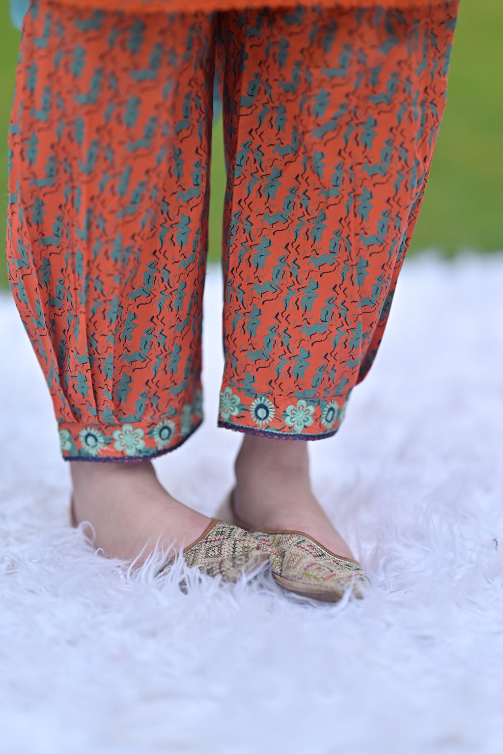 Close-up of orange patterned pants with floral trim worn with beige traditional shoes on a white rug